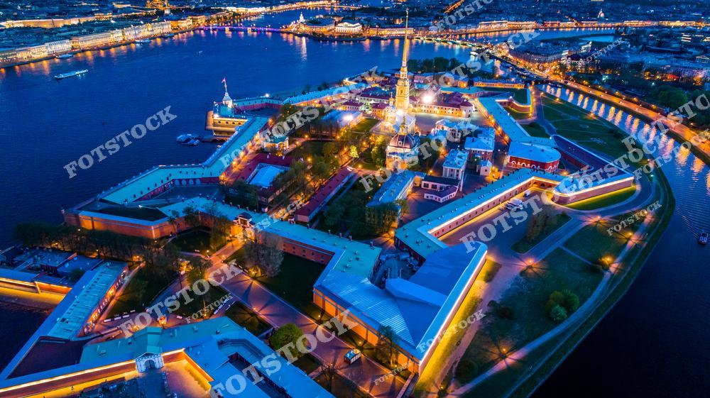 На фотографии изображена Петропавловская крепость, символ основания Санкт-Петербурга. Ночная подсветка подчёркивает геометрические очертания стен и архитектуру центрального собора. На фоне видна река Нева и огни набережной. Впечатляющий вид с высоты передаёт атмосферу исторического центра города.